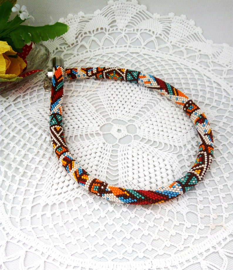 May include: A colorful beaded necklace with a geometric pattern in shades of red, orange, blue, white, and brown. The necklace is made of small beads and has a clasp closure.