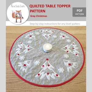 May include: A gray and white quilted table topper with a red border, featuring a snowflake design with Christmas trees and red ornaments. The pattern is called "Gray Christmas" and is a PDF download.