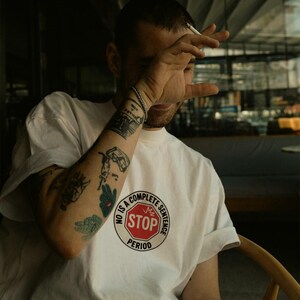 Könnte beinhalten: Weißes T-Shirt mit einem kreisförmigen Aufdruck, der "NO IS A COMPLETE SENTENCE STOP PERIOD" in Schwarz und Rot zeigt. Die Person, die das T-Shirt trägt, hat Tätowierungen auf dem Arm.