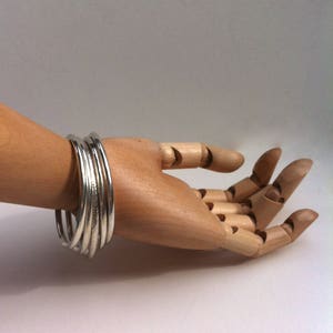 Peut inclure: Plusieurs bracelets argentés empilés sur une main en bois articulée. Les bracelets sont lisses et brillants. La main en bois présente des articulations visibles. L'arrière-plan est de couleur claire et unie.