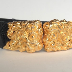 May include: Black belt with gold-colored ornate buckle. The buckle features a swirling, scroll-like design. The belt is made of a textured material and has a wide band. The buckle is a prominent feature.