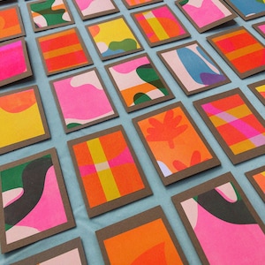 May include: A collection of colourful abstract patterned cards with brown frames. The cards are arranged in rows on a light blue surface.