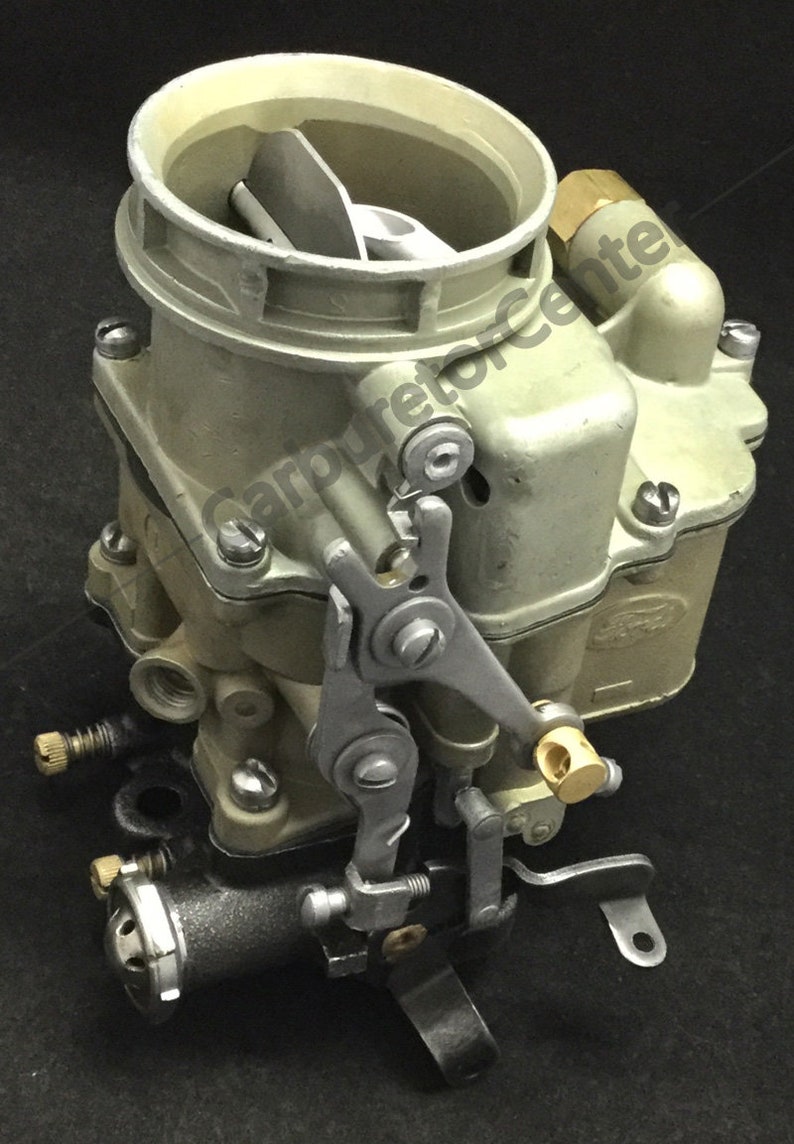 Puede incluir: Un carburador de estilo vintage con un cuerpo de metal plateado y un enlace de acelerador de lat&oacute;n. El carburador tiene una entrada de aire redonda con una cubierta de metal plateado. El carburador est&aacute; montado en una base de metal negro.