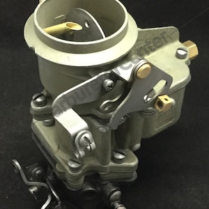 Puede incluir: Un primer plano de un carburador de metal gris con una palanca de latón y una base de metal negro. El carburador tiene una abertura redonda en la parte superior y una serie de tornillos y pernos.