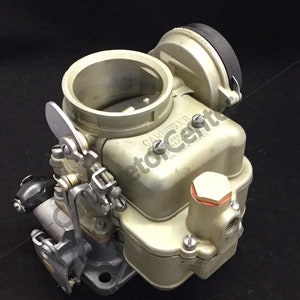 May include: A vintage carburetor with a silver finish. The carburetor has multiple components, including a round air intake, a fuel bowl, and various linkages. The text "CARBURETER" is visible on the body. The item is likely for a classic car or engine.
