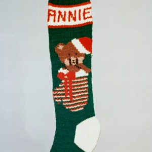 May include: A green Christmas stocking with the name "ANNIE" in red letters. It features a brown teddy bear wearing a Santa hat and a red and white striped pocket. The toe and cuff are white.