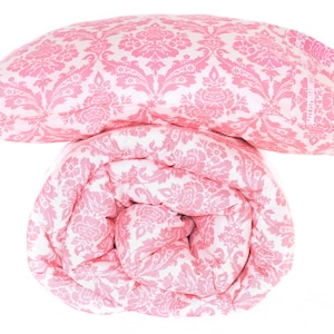 May include: A pink and white damask patterned bedding set, featuring a pillow and a rolled-up comforter. The floral design is repeated across both items. The set is made of a soft, comfortable material, perfect for a bedroom.