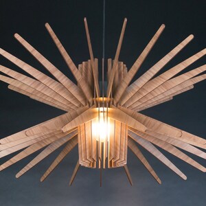 K&ouml;nnte beinhalten: Eine Deckenleuchte aus Holz mit einem Sternburst-Design. Die Leuchte besteht aus d&uuml;nnen, hellen Holzlamellen, die von einem zentralen Punkt nach au&szlig;en strahlen. Im Zentrum der Leuchte ist eine Gl&uuml;hbirne sichtbar.