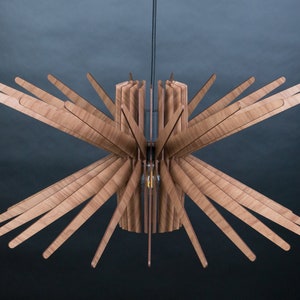 May include: A wooden ceiling light fixture with a starburst design. The light fixture is made of thin, light brown wooden slats that radiate outwards from a central cylinder. The light fixture is suspended from the ceiling by a black cord.
