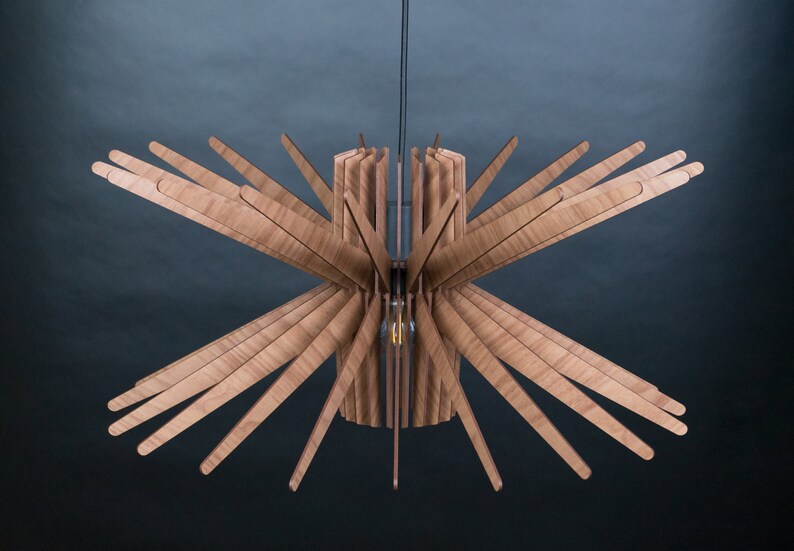 May include: A wooden ceiling light fixture with a starburst design. The light fixture is made of thin, light brown wooden slats that radiate outwards from a central cylinder. The light fixture is suspended from the ceiling by a black cord.