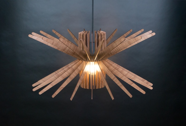 May include: A wooden ceiling light fixture with a starburst design. The light fixture is made of thin, light brown wooden slats that radiate outward from a central cylinder. The light fixture is illuminated by a single light bulb in the center.