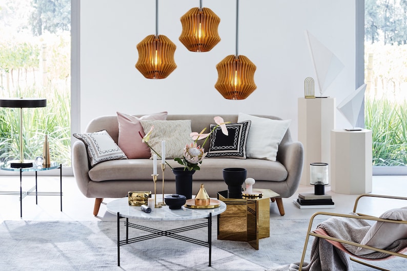May include: A living room interior design featuring a grey sofa, three wooden pendant lights, a coffee table with a marble top, and a gold side table. The room is decorated with throw pillows, a vase with a protea flower, and a gold metal sculpture.