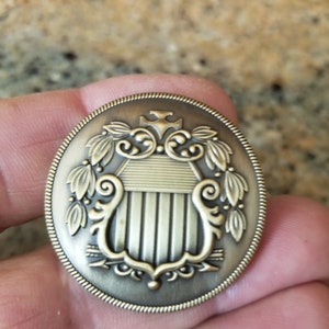 May include: A gold-toned metal pin with an oval shape and a detailed design. The design features a shield with vertical stripes, surrounded by ornate scrolls and leaves.