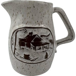 May include: A speckled, off-white ceramic pitcher with a dark brown, oval-shaped relief design. The design depicts a cabin in a winter landscape. The pitcher has a curved handle and a spout for pouring.