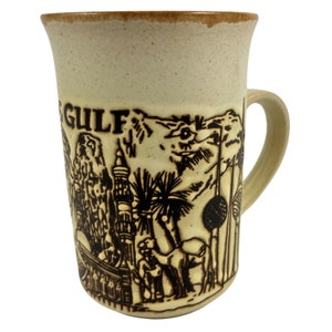 May include: Cream-colored ceramic mug with a brown rim and handle. The mug features a detailed embossed design of a desert scene with the word "GULF" at the top. The design includes buildings, palm trees, and camels.