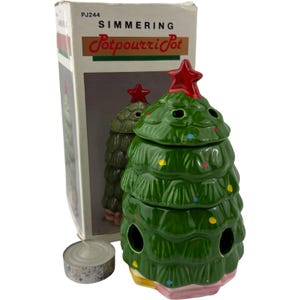 May include: A green ceramic Christmas tree-shaped simmering potpourri pot with a red star topper, colourful painted dots, and ventilation holes. The item includes its original white cardboard packaging labelled Simmering Potpourri Pot and a small tea light candle.