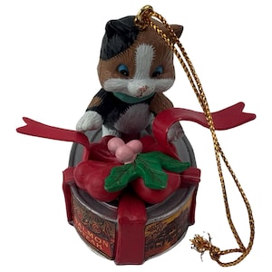 May include: A ceramic cat ornament with a calico pattern, perched atop a red-ribboned gift box. The box is a tin of "Salmon Steak" with a gold string for hanging. The cat has blue eyes and a green collar. A festive holiday decoration.