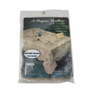 May include: A Progress Creation stamped tablecloth in a clear plastic package. The tablecloth is beige with blue floral embroidery. The package includes 4 napkins and features the text "Royal Mouliné Embroidery Thread Included."