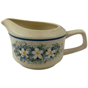 May include: A cream-colored ceramic creamer with a speckled finish, featuring a horizontal dark blue band near the rim and a floral pattern of white daisies with yellow centers surrounded by green and blue foliage on the front side.
