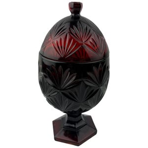 May include: A ruby red, lidded glass jar with a decorative, cut-glass pattern. The jar is egg-shaped and sits on a square pedestal base. The lid has a small, rounded knob. The cut-glass design features a repeating leaf pattern.