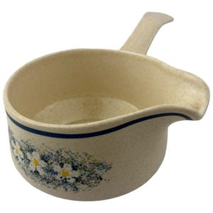 May include: A cream-colored speckled ceramic sauce boat with a single handle and a pouring spout. The exterior features a horizontal dark blue line near the rim and a floral pattern consisting of white daisies with yellow centers and blue foliage.