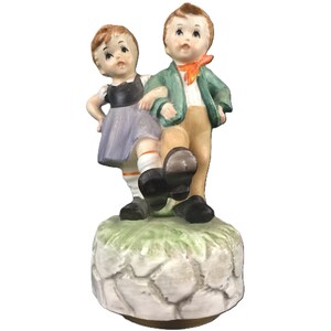 May include: A vintage porcelain figurine of two children dancing. The boy wears a green jacket, orange scarf, and tan pants. The girl has a black top and a lavender skirt. The figurine is mounted on a stone-like base with green grass.