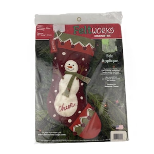 May include: Felt applique kit for a snowman Christmas stocking. The stocking is burgundy with a white snowman wearing a green scarf. The word "Cheer" is written on the stocking. The package is approximately 18 inches long.