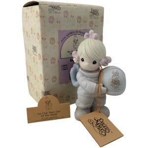 Può includere: Una statuina Precious Moments di un bambino astronauta che tiene un casco. La statuina è di colore chiaro con accenti rosa e si trova accanto alla sua scatola originale. La scatola presenta un motivo ripetuto di figure cherubiche.