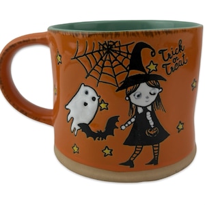 Op de afbeelding: Oranje keramische mok met een Halloween-thema. De mok heeft een heks, een spook, een vleermuis, een spinnenweb en sterren. De woorden "Trick or Treat" staan op de zijkant. Het handvat en de rand zijn bruin en de binnenkant is groen.