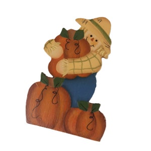 May include: A wooden cutout decoration featuring a scarecrow wearing a straw hat and a green plaid shirt, holding a large orange pumpkin. Two additional orange pumpkins with green leaves and black vine details are positioned at the base.