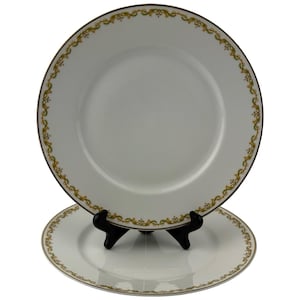 Könnte beinhalten: Zwei weiße Keramikteller mit einem gold-braunen Zierrand. Die Teller werden auf einem schwarzen Tellerständer präsentiert. Die Teller haben ein schlichtes, elegantes Design, das sich für formelle Anlässe eignet.