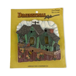 Puede incluir: Adorno de casa de estilo dimensional pintado a mano. La casa es verde con un tejado marrón, una chimenea roja y un acento blanco en forma de nube. El adorno presenta una valla con rosas y una mariposa. El embalaje dice "American Tack & Hardware Co."
