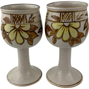 Op de afbeelding: Twee keramische wijnglazen. Elk glas heeft een crèmekleurige voet en steel. De kelk van elk glas is versierd met een gele bloem en bruine bladeren. De rand heeft een bruin geometrisch patroon. De glazen zijn handgemaakt.