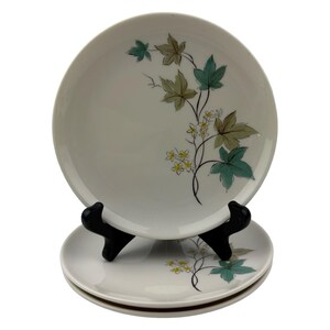 May include: A set of three round white ceramic plates featuring a botanical illustration of green and tan maple-like leaves with small yellow flowers on thin black stems. The plates are stacked and displayed on a black plastic stand.