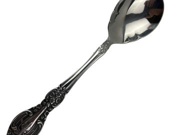 May include: A polished silver-tone metal spoon featuring an ornate handle with embossed floral and scrollwork detailing. The bowl of the spoon is smooth and reflective, showing a clear metallic finish with a curved, rounded shape.