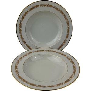 May include: Two white ceramic soup bowls featuring a wide rim decorated with a repeating gold leaf pattern. Each bowl has a thin gold band along the outer edge and the inner rim, providing a consistent metallic accent against the white surface.