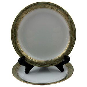 Op de afbeelding: Twee ronde witte porseleinen borden met een brede saliegroene rand versierd met een herhalend gouden bladpatroon. De borden hebben een dunne gouden rand langs de omtrek en staan op een zwarte kunststof standaard tegen een witte achtergrond.
