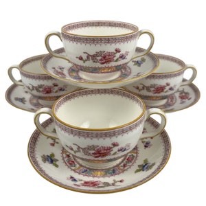 May include: A set of three cream-colored porcelain bouillon cups and saucers featuring a delicate floral pattern in shades of pink, blue, and green, accented with a geometric border and thin gold rims on the edges of the cups and saucers.