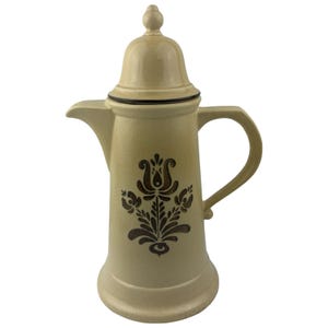 May include: A tall, beige ceramic coffee pot with a matching lid featuring a finial top. The body of the pot is decorated with a dark brown, symmetrical floral pattern. It includes a curved handle on the right side and a spout on the left side.