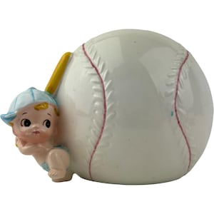 May include: A ceramic baseball figurine featuring a white baseball with red stitching and a small, light-skinned baby figure wearing a blue cap and diaper. A yellow baseball bat is positioned behind the baby.