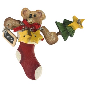May include: A wooden Christmas ornament featuring a brown bear emerging from a red and white stocking. The bear wears a red ribbon and holds a small green Christmas tree with a yellow star, two yellow star charms, and a black sign reading JOY.