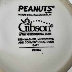May include: White ceramic mug featuring the "PEANUTS" logo and "Gibson" branding. The mug is dishwasher, microwave, and oven safe. The text "WWW.GIBSONUSA.COM" is also visible.