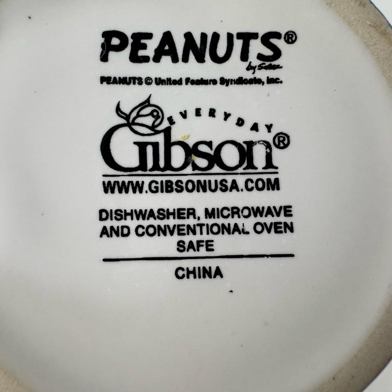 May include: White ceramic mug featuring the "PEANUTS" logo and "Gibson" branding. The mug is dishwasher, microwave, and oven safe. The text "WWW.GIBSONUSA.COM" is also visible.