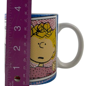 May include: A ceramic mug with a comic-style illustration of a character with yellow hair and a pink dress. The mug is approximately 10 cm tall. The mug has a white handle and a blue rim with a dotted pattern.