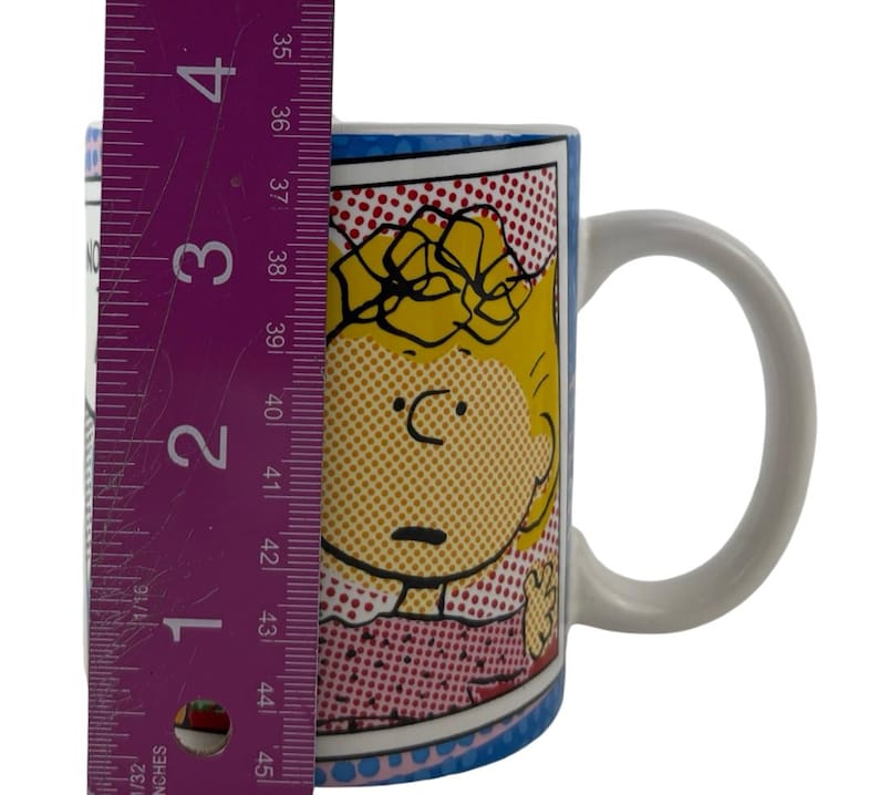 May include: A ceramic mug with a comic-style illustration of a character with yellow hair and a pink dress. The mug is approximately 10 cm tall. The mug has a white handle and a blue rim with a dotted pattern.