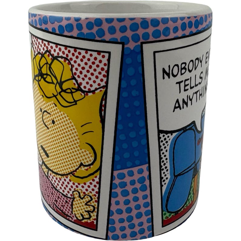 May include: A ceramic coffee mug with a comic book style design. The mug features a cartoon character and the text "NOBODY EVER TELLS ME ANYTHING". The mug has a blue and pink polka dot pattern.