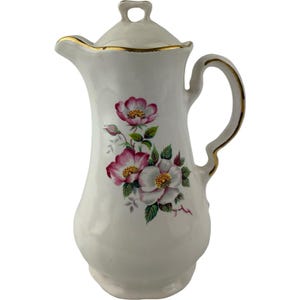 May include: A white porcelain pitcher with a lid, featuring a floral pattern of pink and white wild roses with green leaves. The pitcher has a curved handle and a spout, both accented with a thin gold trim along the edges and the lid handle.