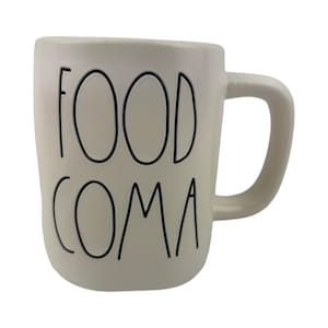 May include: Cream-colored ceramic mug with the words "FOOD COMA" in black, handwritten-style lettering. The mug has a rounded rectangular shape with a curved handle on the right side. The mug is designed for hot beverages.
