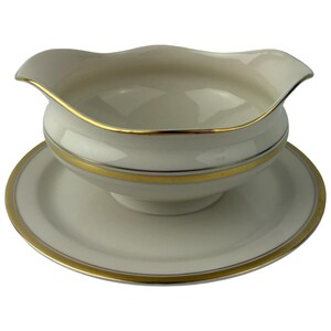 May include: Cream-colored gravy boat with a matching saucer. The boat has a scalloped rim and a gold band around the middle. The saucer also has a gold rim. A classic addition to any dining table.