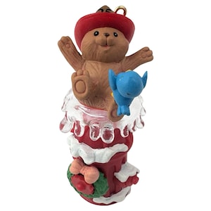 May include: A Christmas ornament featuring a brown bear wearing a red hat, emerging from a red and white stocking. A blue bird sits on the bear's lap. The stocking has a green wreath with a red bow.